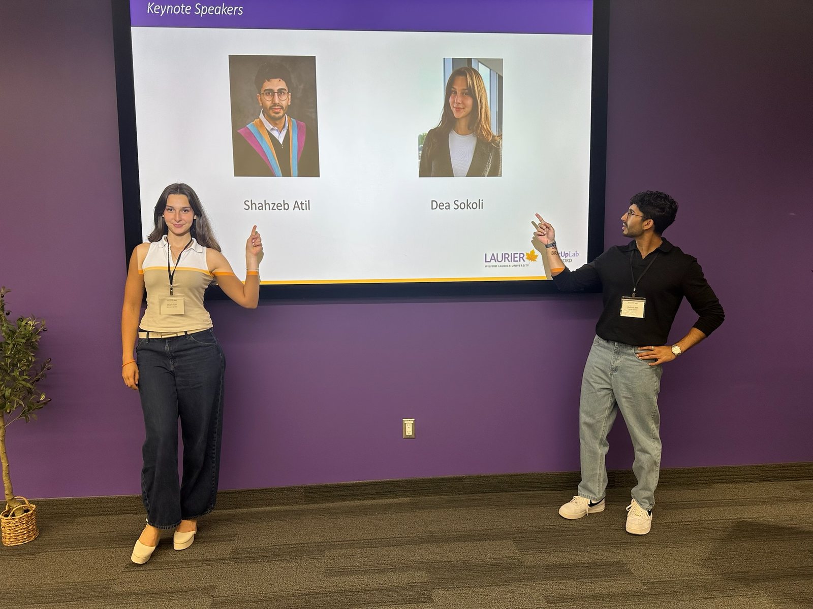 Pointing up at a Keynote Speakers slide at Laurier that shows Shahzeb and Dea's photos and names